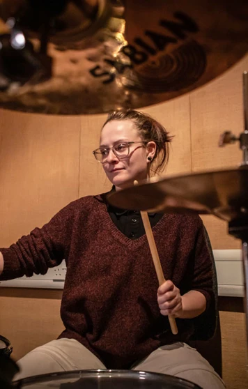 A young drummer with glasses and a burgundy sweater, playing a drum kit surrounded by cymbals. A young drummer with glasses and a burgundy sweater, playing a drum kit surrounded by cymbals.